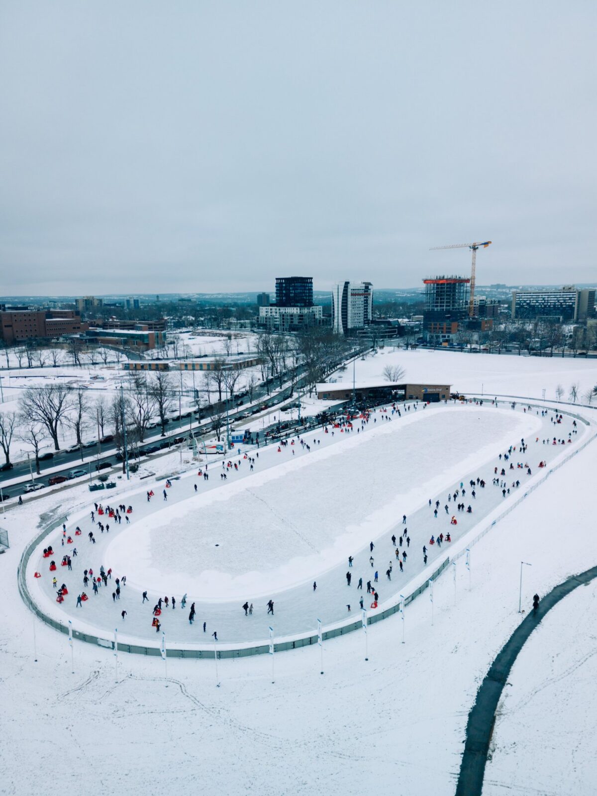 An aerial view of people skating around the Emera Oval on a snowy winter day, with Halifax buildings and construction cranes in the background.