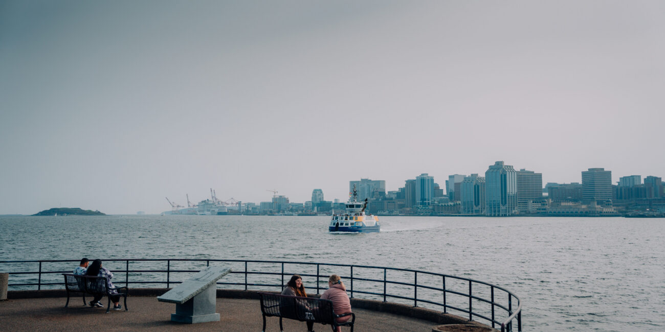 Halifax ferry travelling from Downtown Halifax across the harbour, viewed from a waterfront lookout with benches and people in the foreground.