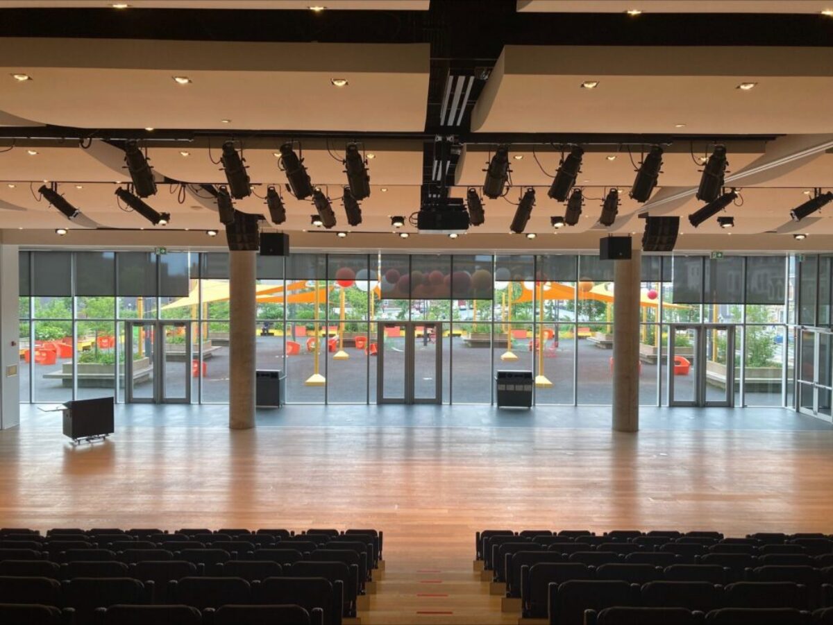 Paul O’Regan Hall at Halifax Central Library set up with rows of seats facing a large stage, with lighting overhead and glass doors opening to the outdoor plaza.